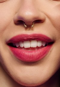 Bouche souriante avec un rouge à lèvres rouge vif, des dents blanches et un piercing septum doré sur un teint clair.