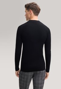 Uomo con capelli corti e ricci, che indossa un maglione nero a coste con maniche lunghe e pantaloni grigi a scacchi, girato di spalle su uno sfondo neutro.