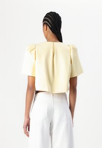 Hoch geschnittenes gelbes Oberteil mit puffärmeligen kurzen Ärmeln und einer Rückenfalte, kombiniert mit hoch geschnittenen weißen Hosen. Glatte Textur, minimalistisches Design.