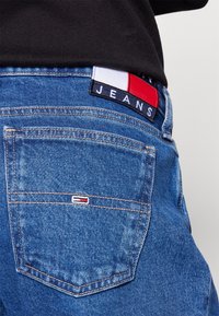 Blue denim jeans with a back pocket featuring a small logo tag. The waistband has a colorful patch displaying "JEANS" and a flag design.