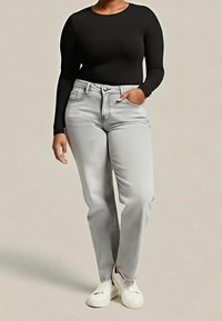 Personne portant un haut noir ajusté à manches longues, un jean taille haute gris clair, des baskets blanches, avec une main dans la poche, sur un fond uni.