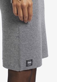 Main posée sur une jupe en tricot gris avec une étiquette rectangulaire noire de la marque Khujo près de l'ourlet, au-dessus de collants foncés.