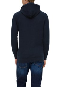 Marineblaue Strickkapuzenjacke mit gerippter Textur, einer Kordelzugkapuze und langen Ärmeln. Über dunkelblauen Jeans getragen, zeigt die Rückansicht.