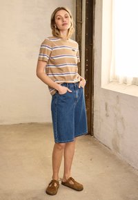 Femme debout à l'intérieur près d'une fenêtre, portant un t-shirt beige rayé, un short en jean bleu jusqu'aux genoux et des chaussures marron sans lacets.