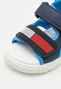 Sininen sandaali tummansinisillä hihnoilla ja kumitossupäällä. Valkoiset ja punaiset korostukset hihnassa. Teksturoitu kangas ja joustava pohja.