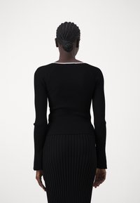 Kvinna med flätat hår i en knut, klädd i en svart ribbad långärmad topp och matchande ribbad kjol, vänd bort mot en vit bakgrund.