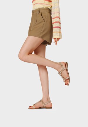 Gambe che indossano pantaloncini marroni a vita alta e sandali piatti dorati con gioielli, con una maglietta a maniche lunghe a righe, una gamba leggermente sollevata all’indietro.