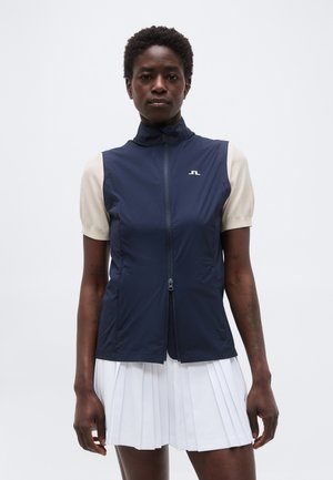 Žena oblečená v tmavomodrej bezrukávovej veste na zips, cez béžové tričko s krátkym rukávom a biele plisované sukni, stojaca pred jednoduchým svetlým pozadím.