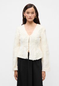 Biela textúrovaná blúzka s vlnkovými vzormi, V-výstrihom a predným zapínaním na gombíky. Dlho, priehľadné rukávy a priliehavý strih.