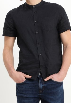 Männlich, trägt ein schwarzes kurzärmliges Hemd mit Stehkragen und Knöpfen, die Hände in die Taschen einer dunkelblauen Jeans gesteckt.