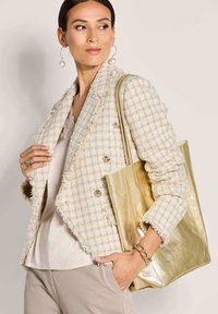 Beige ruiten tweed blazer met rafelige randen, gecombineerd met een metalen gouden handtas. Zacht crèmekleurig topje en lichtbeige broek.