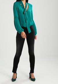 Kvinde iført en teal langærmet bluse med halsbånd, sorte skinny jeans og teal højhælede sko, stående med afslappet holdning.
