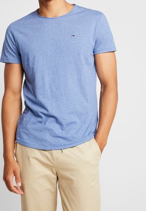 Mężczyzna w jasnoniebieskiej koszulce z krótkim rękawem i beżowych spodniach, z jedną ręką w kieszeni na jednolitym tle.