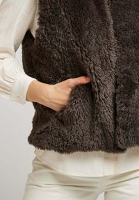 Gilet en fausse fourrure marron avec une finition douce et texturée, doté de poches latérales et d'une coupe décontractée. Porté par-dessus une chemise en tissu léger.