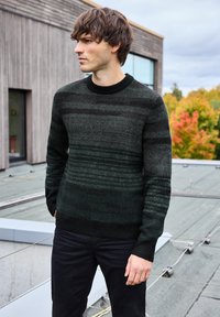 Mörkgrön randig stickad tröja med ribbad halsringning. I kombination med svarta byxor står modellen utomhus framför en trästruktur.