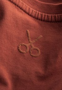 Roestkleurige stof met een zachte textuur, met een geborduurd paar scharen in een donkerdere tint, gepositioneerd in het midden.