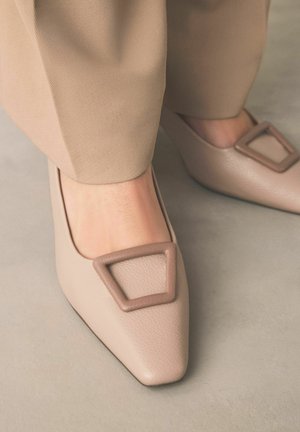 Smėlio spalvos butai su smailiais noseliais, pagaminti iš tekstūruotos odos, papuošti didelėmis geometrinėmis sagtimis, dėvimi kartu su derančiomis smėlio spalvos kelnėmis ant pilko grindinio.