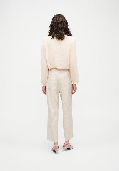 Femme aux cheveux bouclés, vue de dos, portant un chemisier léger beige à manches longues, un pantalon taille haute assorti et des sandales à talons bas.