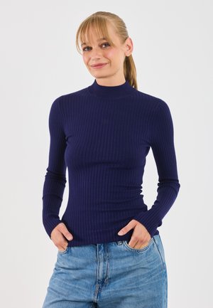 Kvinne med blond hestehale iført en marineblå ribbestrikket genser med lange ermer og blå jeans, står med hendene i lommene mot en hvit bakgrunn.