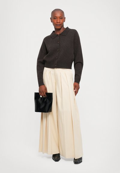 Femme portant un cardigan boutonné marron foncé et un pantalon plissé large crème, tenant un sac à main noir, se tenant devant un fond blanc.