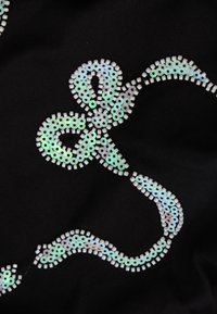 Μαύρος ύφασμα διακοσμημένο με έναν ιριδίζοντα σχεδιασμό από παγιέτες σε σχήμα φιόγκου, με στρογγυλές κα�ι ορθογώνιες παγιέτες που έχουν γυαλιστερή επιφάνεια.