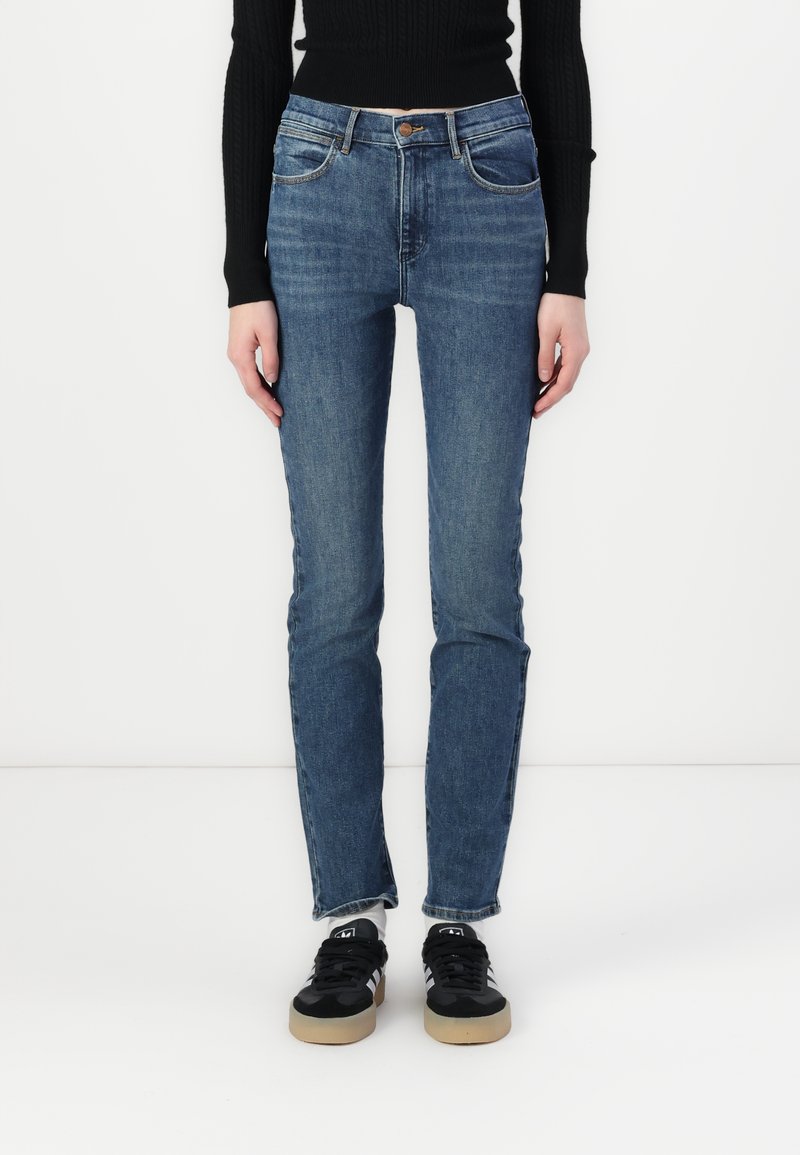 Blue denim jeans with a straight-leg fit, five pockets, a button closure, and subtle faded detailing. Paired with black sneakers.