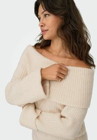 Sweter dzianinowy w kolorze kremowym, z odkrytymi ramionami, o prążkowanej strukturze, szerokich rękawach i wzorze warkoczowym. Miękki, przytulny materiał.