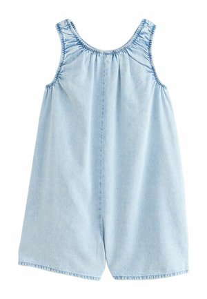 Mladé dievča s dlhými kučeravými vlasmi, ktoré má na sebe svetlomodrý bezrukávový denimový overal a žlté sandále, stojace na béžovom pozadí.