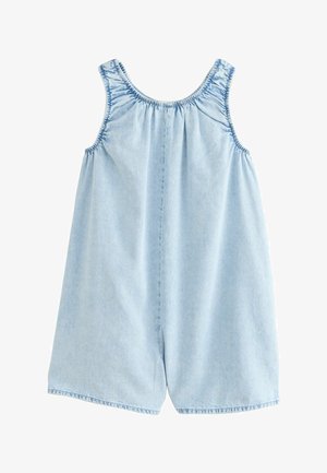 Jeune fille aux longs cheveux bouclés portant une combinaison courte en denim bleu clair sans manches et des sandales jaunes, debout devant un fond beige.