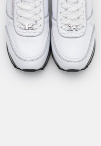 Baskets blanches avec une tige en cuir lisse, des accents argentés et un bout arrondi. Comprend des lacets plats, un détail de logo et une semelle noire texturée.