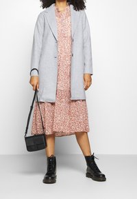 Vrouw in een roze bloemen-midi-jurk, grijze jas, zwarte veterschoenen en met een kleine zwarte handtas tegen een effen achtergrond.