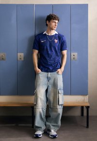 Maglia Nike blu con accenti rosa a contrasto, dotata di un emblema di gallo. Abbinata a jeans cargo leggeri blu chiaro e sneaker bianche.