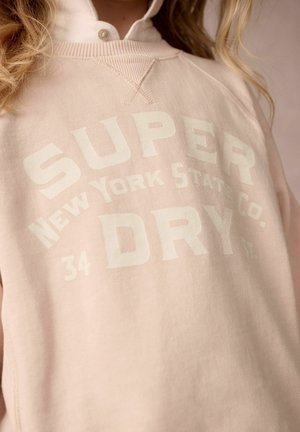 Felpa rosa chiaro con colletto a coste, con testo bianco in grassetto "SUPER DRY" e "New York State Co." sul davanti. Tessuto morbido.