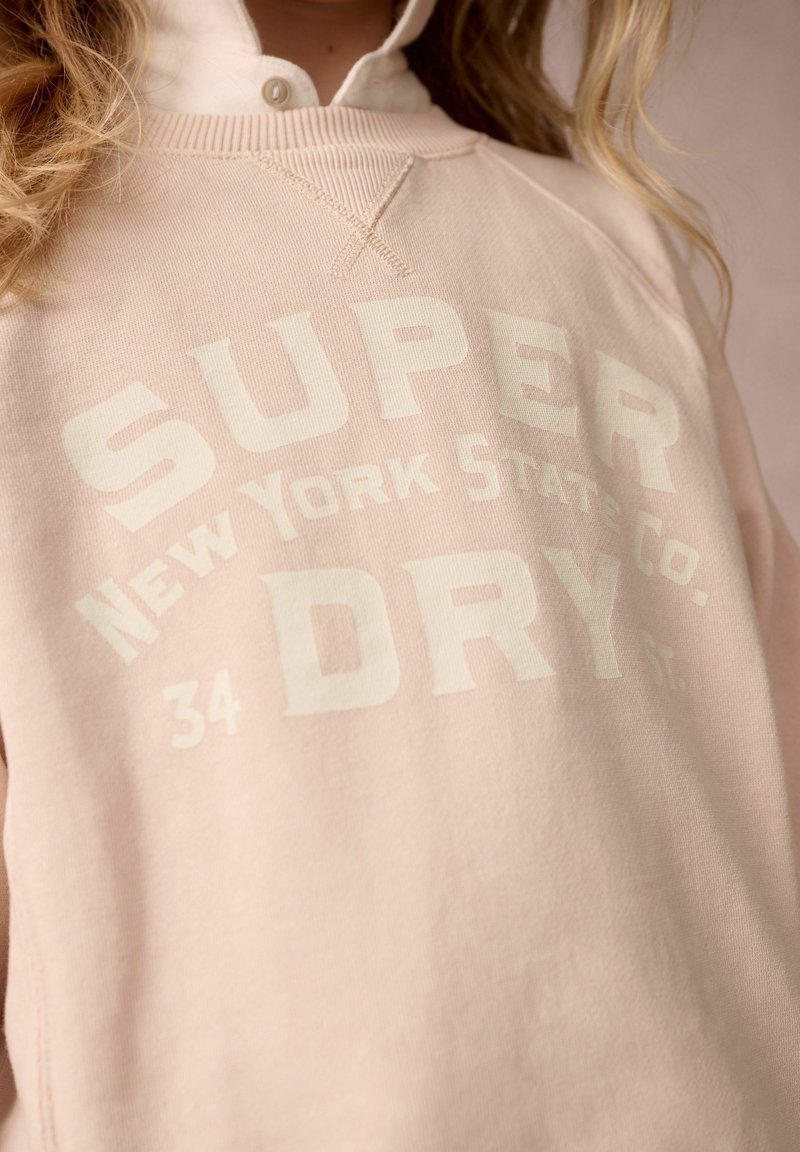 Vaaleanpunainen collegepaita, jossa on ribbejä kauluksessa, etupuolella valkoista rohkeaa tekstiä "SUPER DRY" ja "New York State Co.". Pehmeä kankaan rakenne.