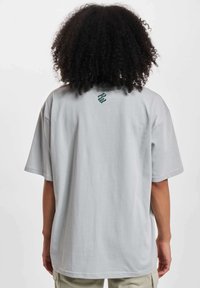Lys grå oversized t-skjorte laget av bomull, med rund hals og korte ermer. En liten grønn logo er plassert på øvre rygg.