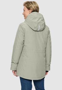 Vrouw in een lichtgrijze gewatteerde winterjas met capuchon, gezien vanuit de achterkant, in combinatie met blauwe jeans tegen een effen lichte achtergrond.