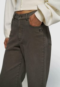 Person trägt dunkelgraue Hose mit "10" Stickerei nahe der rechten Tasche, Hand in der Tasche, und cremefarbenes, gekürztes Sweatshirt.