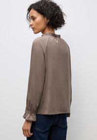 Blusa marrone a maniche lunghe con scollo arricciato e maniche con polsini, realizzata in un tessuto morbido, indossata sopra un paio di jeans in denim blu.