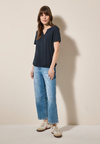 Mörkblå topp med kort ärm och V-ringning som matchas med ljusblå vida jeans och vita sneakers med beige och röda accenter.