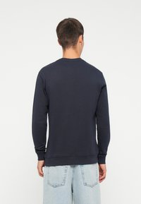 Granatowy sweter z miękkiego materiału, z okrągłym dekoltem, długimi rękawami oraz ściągaczami przy mankietach i dolnym brzegu. Noszony z jasnoniebieskimi dżinsami.
