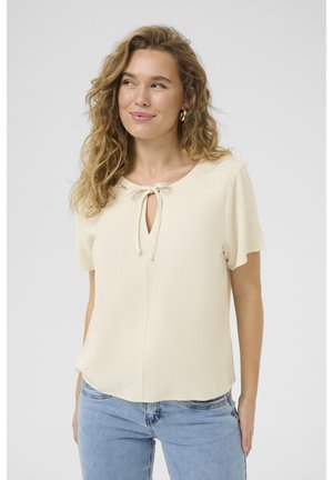Blusa a maniche corte color crema con scollo rotondo e dettaglio con fiocco nella parte anteriore. Realizzata in tessuto leggero e texturizzato.