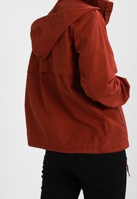 Chaqueta ligera roja con capucha, de corte holgado, con un diseño de solapa en la espalda y un bolsillo con cremallera en el lateral.