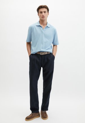 Mężczyzna z rękami w kieszeniach, ubrany w jasnoniebieskie polo z krótkim rękawem, ciemnogranatowe spodnie, beżowy pasek i brązowe zamszowe buty na białym tle.