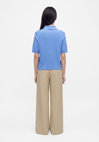 Femme aux longs cheveux noirs portant un polo bleu clair à manches courtes et un pantalon large beige, debout de dos sur un fond blanc.