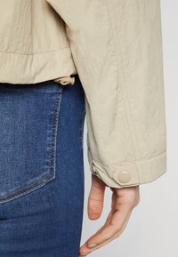 Nærbillede af en person iført blå jeans og en beige jakke med en knappet manchet, hånden afslappet ved deres side.