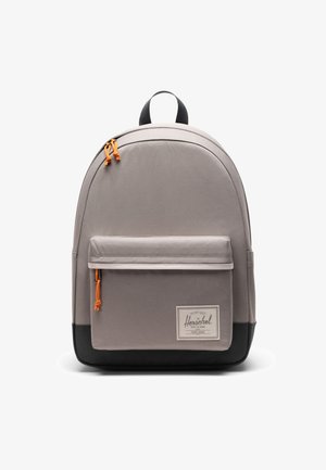 Mochila de tela gris con forma curva, base negra, bolsillo frontal y acentos de tiradores de cremallera color naranja. Presenta un asa superior y una parche de logo.