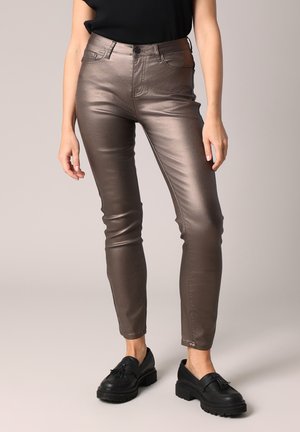 Jeans en bronze métallique avec une coupe ajustée, taille haute, léger éclat et cinq poches, associés à des mocassins noirs.
