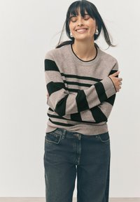 Pull en maille gris avec des rayures noires, col rond et poignets côtelés. Porté avec un jean en denim noir, présentant une coupe décontractée.