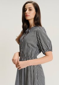 Donna con lunghi capelli castani che indossa un vestito a scacchi bianco e nero con maniche a sbuffo, in piedi su uno sfondo chiaro e neutro.