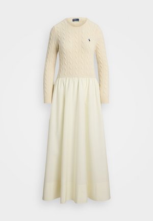 Robe en tricot torsadé crème avec manches longues, col rond et jupe flottante plissée, comportant un petit logo foncé sur la poitrine.